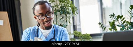 Fröhlicher afroamerikanischer Arzt mit Pillen in der Hand, der auf Laptop-Kamera, Telemedizin, Banner schaut Stockfoto