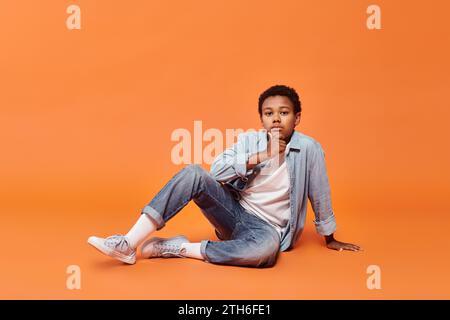 Nachdenklicher junger afroamerikanischer Junge in lässiger Kleidung, der auf dem Boden sitzt und in die Kamera schaut Stockfoto