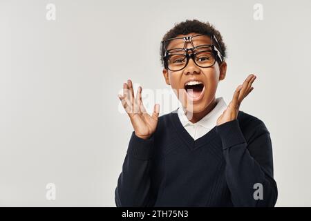 Aufgeregtes afroamerikanisches Kind in Pullover und Brille mit Brille und Blick auf die Kamera auf Grau Stockfoto