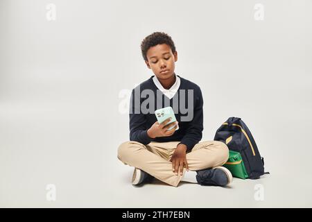 afroamerikanischer Schuljunge in Uniform mit Smartphone und in der Nähe des Rucksacks auf grauem Hintergrund Stockfoto