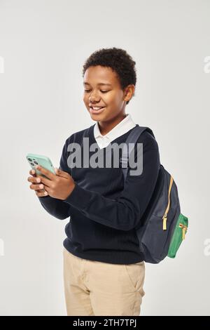 Glücklicher afroamerikanischer Schuljunge in Uniform mit Smartphone und auf grauem Hintergrund stehend Stockfoto