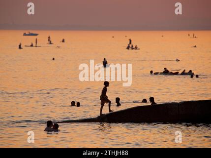 Silhouetten von Menschen, die den Strand bei Sonnenuntergang, Brighton & Hove genießen. Der Himmel erleuchtet mit dem schwindenden Licht des Tages. Boot- und Paddelboarder auf dem Meer in der Ferne. England, Großbritannien Stockfoto