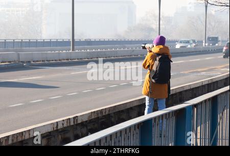 Eine Straßenfotografin, die auf der Brücke steht und mit der Kamera fotografiert, an einem verschmutzten nebeligen Tag, von hinten Stockfoto
