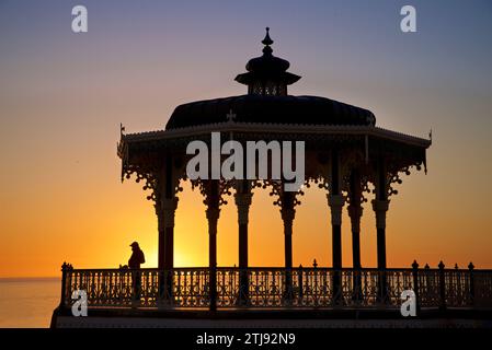 Silhouettenform von Brightons viktorianischem Bandstand mit einem einsamen Mann bei Sonnenuntergang. Brighton & Hove, East Sussex, England. Blauer und goldener Himmel. Stockfoto