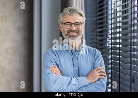 Porträt eines reiferen, erfolgreichen Geschäftsmanns Boss, eines älteren grauhaarigen Mannes im Büro, der mit überkreuzten Armen in die Kamera blickt, ein erfahrener Bankkaufmann im Hemd. Stockfoto
