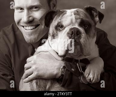 Und ein schwarz-weißes Porträt eines Mannes, der seine Haustier-Bulldogge hält und in die Kamera schaut Stockfoto
