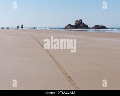 Ein paar Leute laufen an einem Sandstrand mit Felsvorsprüngen und Quad-Bikes dahinter in der Stadt Essaouira, der „windigen Stadt“, Marokko. Dezember 2023 Stockfoto