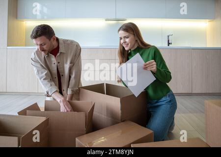 Glückliche verheiratete Hausbesitzer, die in eigene neue Wohnungen ziehen, Kartons auspacken und die Dinge mit einer Liste überprüfen Stockfoto