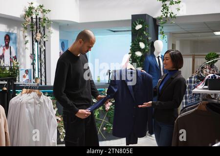 Shoppingcenter-Boutique-Frau, die Jacke am Kleiderbügel hält und arabischen Kunden bei der Wahl des Outfits hilft. Bekleidungsgeschäft asiatische Mitarbeiter beraten Kunden, helfen beim Kauf Stockfoto