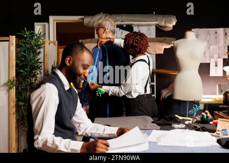 Teamwork-Couturiers helfen älteren Kunden, perfekt passende maßgeschneiderte Business-Anzüge zu finden. Glückliche Schneider, die bei der Auftragsvergabe für kundenspezifische Anpassungen Messungen vornehmen Stockfoto
