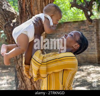 afrikanische Mutter mit ihrem Baby, das ihn in der Luft über ihrem Kopf hält, spielen sie draußen im Hof Stockfoto