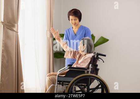 Ältere asiatische Frau, die mit Physiotherapeuten im Wohnzimmer im Rollstuhl Dehnübungen macht. Stockfoto