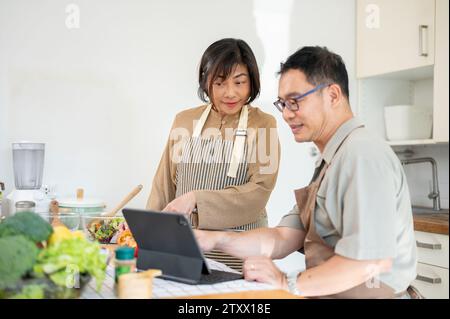 Schöne, glückliche Erwachsene asiatische Paare schauen sich ein Online-Rezept auf einem digitalen Tablet an und kochen gemeinsam in der Küche. Familienverbundenheit, Hauskochen Stockfoto