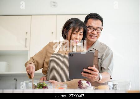 Schöne, glückliche Erwachsene asiatische Paare schauen sich ein Online-Rezept auf einem digitalen Tablet an und kochen gemeinsam in der Küche. Familienverbundenheit, Hauskochen Stockfoto