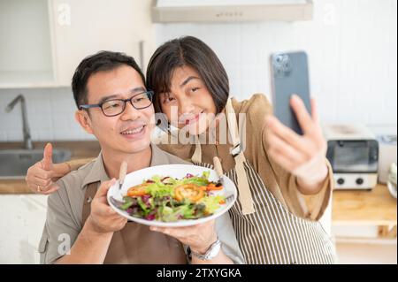 Ein hübsches asiatisches Paar, das Fotos oder Selfies mit ihrem Essen in der Küche macht. Familie, Ehemann und Ehefrau Stockfoto