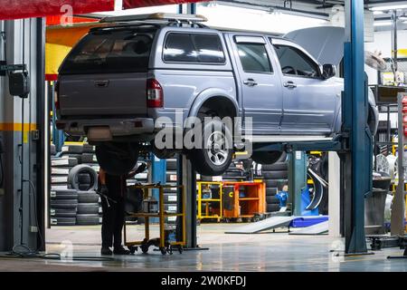 Der Pickup-Truck wird in einer Servicewerkstatt mit einem mechanischen Brückenheber in die Luft gehoben, um bei Nacht Reparaturen und Wartungsarbeiten durchzuführen, Rückansicht. Stockfoto