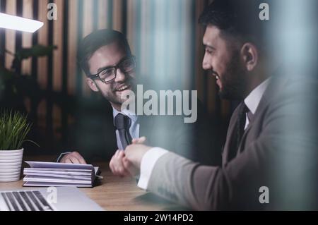 Zwei positive, selbstbewusste Geschäftsmänner, die nach der Verhandlung die Hände über den Besprechungstisch schütteln, einen Deal abschließen, einen Vertrag abschließen, eine Vereinbarung treffen, lächeln. Stockfoto