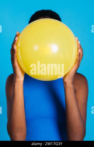 Frau, die einen gelben Ballon vor blauem Hintergrund in einem Studio bläst. Frau mit kurzen Haaren, die einen gelben Ballon aufbläht. Stockfoto
