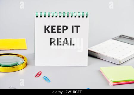 HALTEN SIE ES ECHT, die Inschrift auf dem Notizbuch auf grauem Hintergrund neben dem Briefpapier Stockfoto