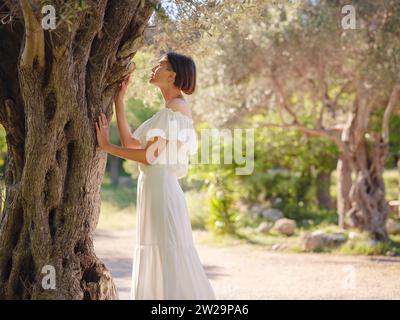 Schöne asiatische junge Frau in weißem Kleid draußen in der Nähe von Olivenbäumen. Genießen Sie frische Luft und nehmen Sie an Outdoor-Aktivitäten Teil. Friluftsliv Konzept bedeutet, so viel Zeit wie möglich im Freien zu verbringen Stockfoto