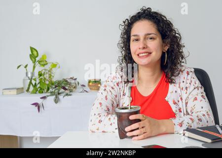 Porträt einer jungen lateinischen Geschäftsfrau mit Lockenwicklern und weiß-roten Freizeitkleidung. Sie sitzt mit ihrem Kollegen an ihrem Schreibtisch und schaut her Stockfoto
