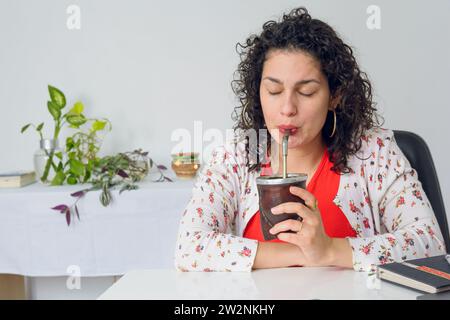 Junge Latina Geschäftsfrau, mit Lockenwicklern und lässigen Kleidern, in ihrem Heimbüro sitzend am Schreibtisch glückliche trinkende Freundin mit geschlossenen Augen, mit Tisch Stockfoto
