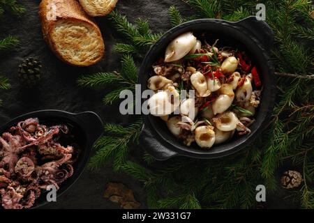 Gebratener Tintenfisch mit Chilischote, Knoblauch und Butter, serviert in einer eisernen Pfanne auf schwarzem, strukturiertem Hintergrund, dekoriert mit Walddetails. Weihnachtsessen Stockfoto