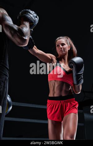 Sportliche Frau in roten Shorts und Oberteil boxt mit einem Trainer. Boxen und Mixed Martial Arts Konzept. Gemischte Medien Stockfoto