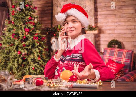 Bild einer schönen Frau in weihnachtlicher Kleidung. Sie spricht am Telefon, während sie Silvestergeschenke in einem Landhaus verpackt. Feiertagskonzept. Gemischt Stockfoto