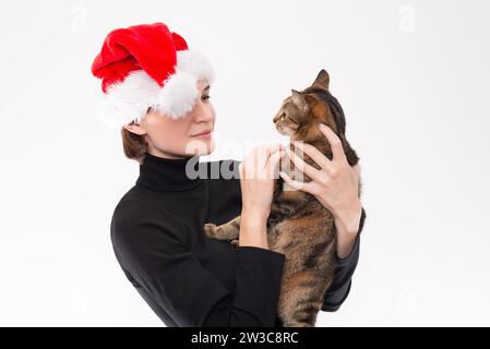 Bild einer Frau mit einem Weihnachtsmann-Hut, die eine Katze im Arm hält. Heller Studiohintergrund. Gemischte Medien Stockfoto