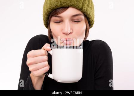 Porträt einer Frau in grünem Strickmütze mit einem Becher in den Händen. Saisonales Viruskrankheitskonzept. Gemischte Medien Stockfoto