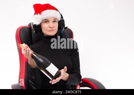 Bild einer Frau mit Weihnachtsmann-Hut, die auf einem Spielstuhl sitzt und Kopfhörer trägt und eine Flasche Champagner zeigt. Heller Studiohintergrund. Gemischt Stockfoto