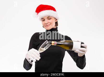 Bild einer Frau in einem Weihnachtsmann-Hut, die Champagner aus einer Flasche gießt. Heller Studiohintergrund. Gemischte Medien Stockfoto