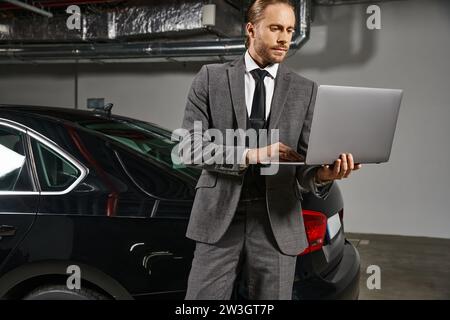 Gut aussehender Mann im schicken Anzug, der an einem Laptop auf dem Parkplatz in der Nähe seines Autos arbeitet, Geschäftskonzept Stockfoto