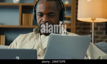 afroamerikanischer Mann Lehrer Geschäftsmann Freiberufler im Headset, der Dokumente überprüft und Daten in Laptop eingibt, der zu Hause mit dem Computer arbeitet Stockfoto
