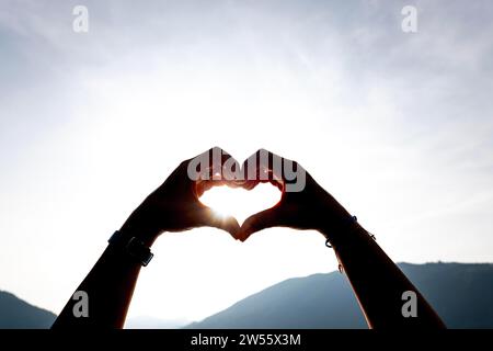 Frauenhände machen eine Herzform gegen den Himmel mit Sonne und Sonnenstrahl und Berg in der Schweiz Stockfoto