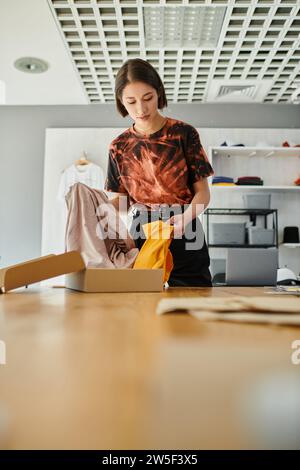 Stilvoller asiatischer Modedesigner, der während seiner Arbeit im Druckstudio den Karton mit Kleidungsstücken auspackt Stockfoto