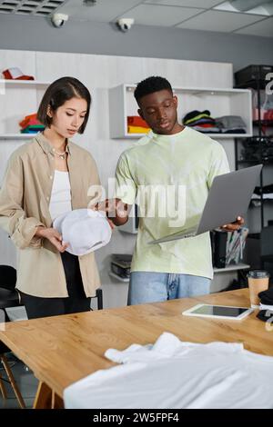 Multiethnische Modedesigner mit Laptop und weißer Kappe diskutieren über neues Projekt im Druckstudio Stockfoto