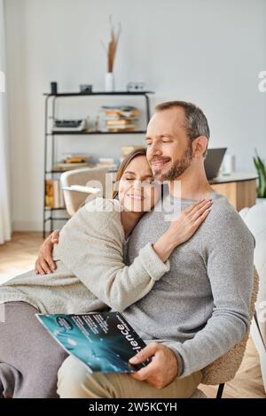 Eine entzückte Frau umarmt Ehemann, sitzt mit dem Wissenschaftsmagazin auf der Couch zu Hause, kinderfreies Leben Stockfoto