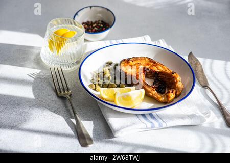 Hoher Winkel von gut zubereitetem, gegrilltem Forellensteak mit Kapern und Zitrone, serviert auf einem weißen Teller mit blauem Rand auf gestreifter Serviette, Besteck und Glas Stockfoto