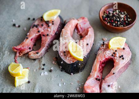 Hoher Winkel frischer Forellensteaks auf Backpapier, fertig zum Kochen mit Zitronenscheiben, Salz und Pfeffer in der Nähe einer kleinen Schüssel gemischter Gärung Stockfoto