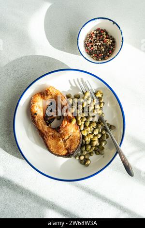 Blick von oben auf gut vorbereitetes gegrilltes Forellensteak mit Kapern, serviert auf einem weißen Teller mit Gabel auf grauer Oberfläche in der Nähe einer Schüssel mit Kapern Stockfoto