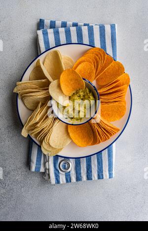 Blick von oben auf den Teller mit verschiedenen Chips, einschließlich Kartoffeln, Paprika und Käse, serviert mit einer Schüssel mit frischer Avocado-Guacamole auf einer gestreiften Serviette Stockfoto