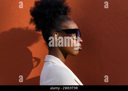 Seitenansicht einer jungen schwarzen Modeunternehmerin mit schwarzem Lockenhaar mit Brille an roter Wand Stockfoto