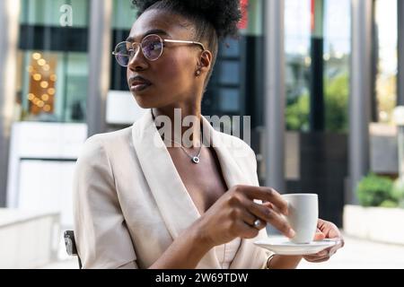 Nachdenklich ruhige afroamerikanische Frau mit Brille und schwarzem Haar, die eine Kaffeetasse hält und wegblickt, während sie im Café sitzt Stockfoto