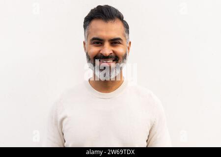 Bärtiger Mann in weißem Langarm, der in die Kamera blickt und lächelt, während er vor schlichtem Hintergrund steht Stockfoto