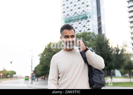 Positiver bärtiger indischer Geschäftsmann mit einem Rucksack, der lächelnd und auf dem Smartphone vor einem verschwommenen Stadtbild spricht Stockfoto