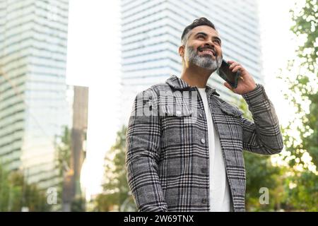 Niedriger Winkel eines positiv bärtigen indischen Geschäftsmannes in karierter Jacke lächelnd und redend auf Smartphone vor verschwommenem Stadtbild Hintergrund Stockfoto