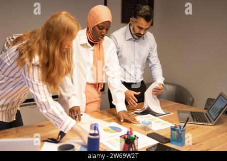 Junge Kollegen aus verschiedenen Bereichen, die sich am Schreibtisch mit Laptop treffen und Dokumente während des Brainstormings in modernen Büros diskutieren Stockfoto