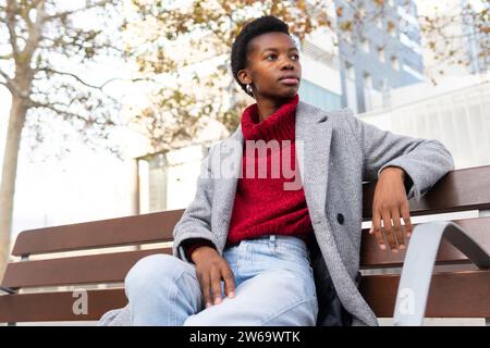 Von unten unemotionale Erwachsene afroamerikanische Frau in roten Pullover Jeans und grauem Mantel, die auf einer Holzbank in der Stadt ruht und das Wochenende genießt Stockfoto